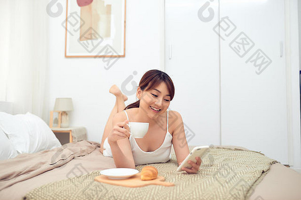 早上躺在床上，兴高采烈的亚洲年轻女子正在使用手机