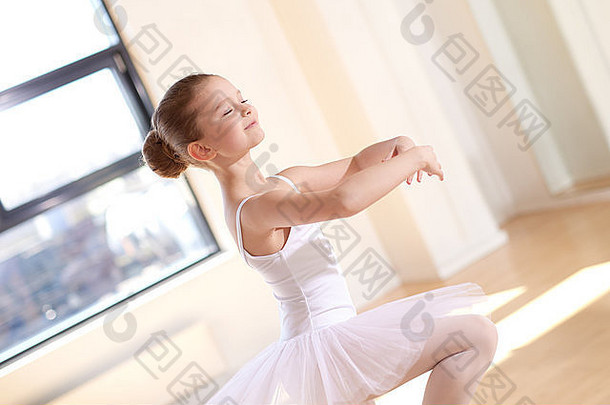 身着白色芭蕾舞裙、面带微笑的美丽芭蕾舞女孩独自在演播室里练习舞蹈。