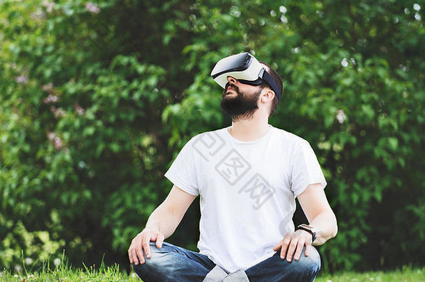 年轻的有胡子的男人。穿虚拟现实护目镜坐着花园生活方式有趣的放松概念