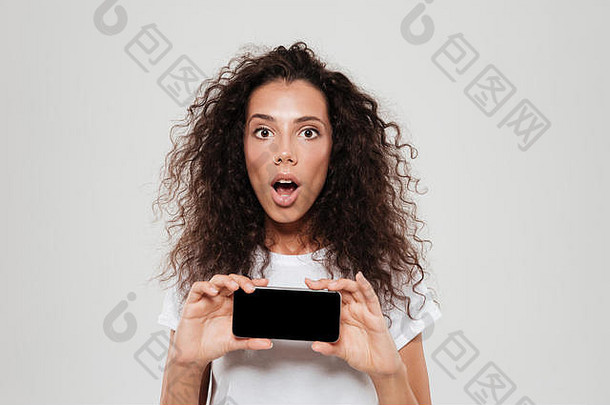 一个惊讶的卷发女人张<strong>大嘴巴</strong>，在灰色背景下看着空白的智能手机屏幕
