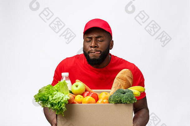 送货概念-英俊的非裔美国人送货员从商店里拿着食品和饮料的包装盒。在灰色工作室背景上隔离。拷贝空间