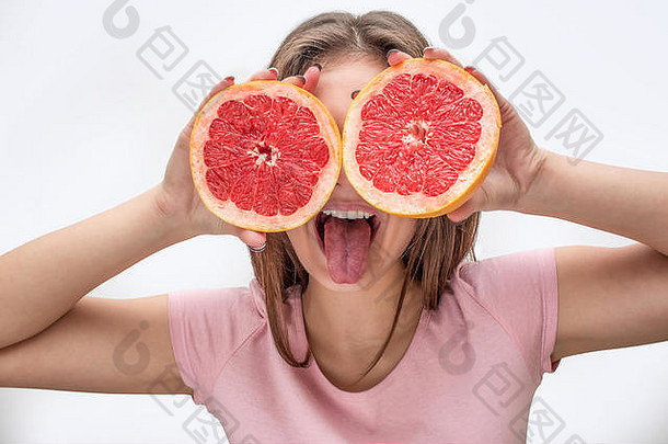 顽皮的年轻女子用两半葡萄柚遮住眼睛。<strong>模特秀</strong>舌头。在灰色背景上隔离。