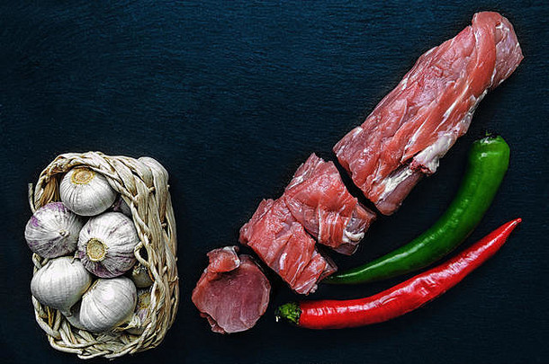 柳条筐里放着新鲜的猪肉片和大蒜，黑色黑板上放着红椒和青椒。水平框架。