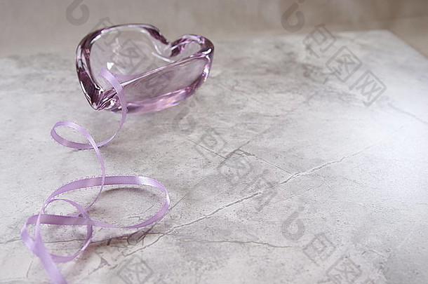柔软的淡紫色彩带卷曲成心形浅紫色，背景为淡紫色玻璃盘。大理石表面。