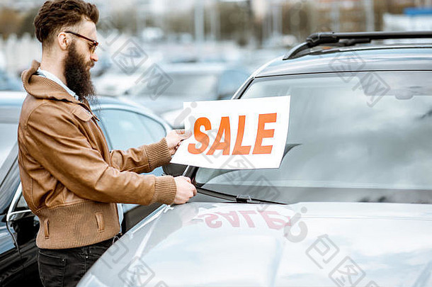 在一家经销店的空地上，一位留着时髦胡须、挂着销售牌照的推销员站在汽车旁边