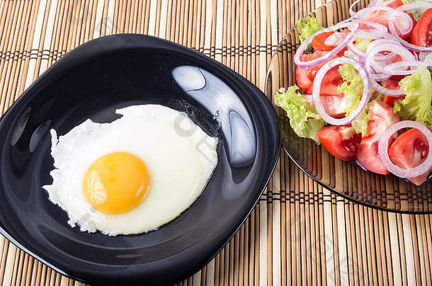 桌上的家常菜——黑盘子上的蛋黄煎蛋和条纹垫子上的西红柿、生菜和洋葱沙拉