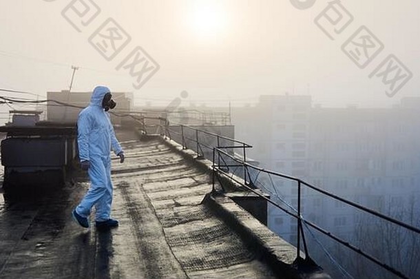 科学家穿着白色防护服和防毒面具，在一座城市建筑的屋顶上行走。背景是初升的太阳和雾天。空间。环境问题的概念