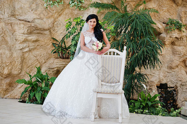 诱惑模特黑发新娘穿着激动人心的婚纱，双手捧着花束坐在白色的椅子旁