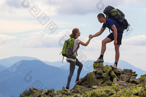 年轻的游客背包运动男孩帮助苗条的女孩气候岩石山前明亮的夏天天空山范围背景touri