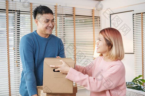这对幸福的亚洲夫妇搬进客厅的新房时拿着纸板箱。为新家打开行李。女人为丈夫擦汗