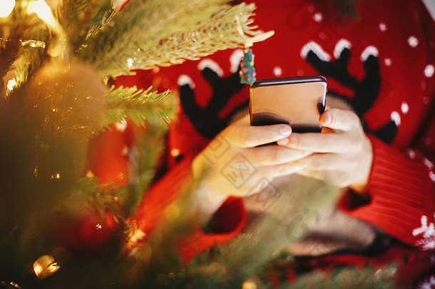 美丽的女孩手持智能手机，在节日房间里浏览金色圣诞树，圣诞树上挂着彩灯和礼物。圣诞节网上购物中心