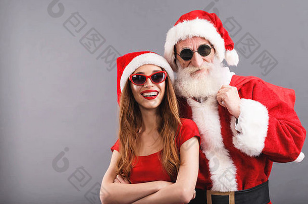 白胡子的圣诞老人戴着太阳镜，年轻的圣诞夫人戴着圣诞帽、红色连衣裙和太阳镜站在灰色背景上微笑着，新年、圣诞节、假日、纪念品、礼品、<strong>购</strong>物、折扣、商店、雪人圣诞老人、化妆、发型、<strong>狂欢</strong>节。