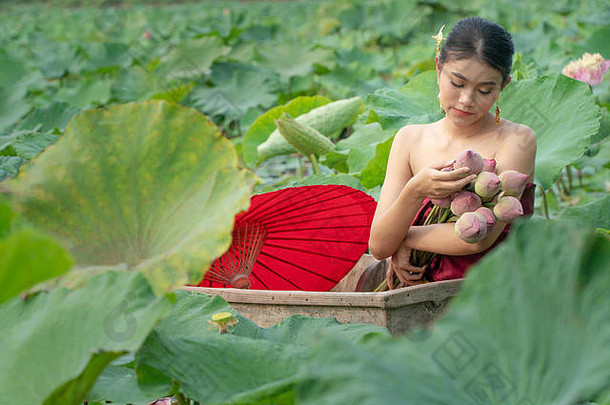 美丽的亚洲妇女穿着传统的泰国服装，坐在木船上，打着红色的纸伞，在莲花湖上。她的手拿着一个p