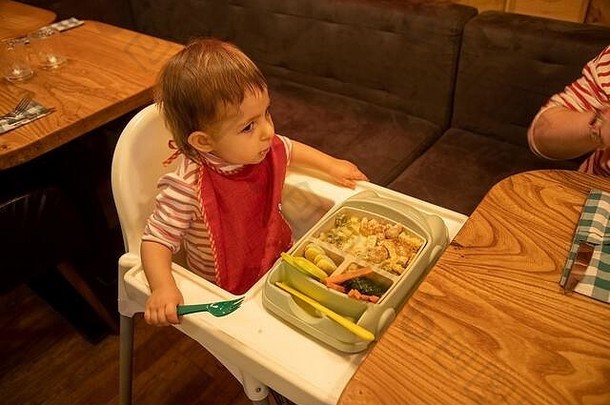 在咖啡馆和餐馆里吃孩子的概念。小可爱的孩子坐在高椅子上，旁边的桌子上放着一盘美味可口的开胃食物
