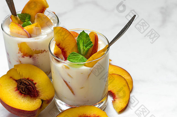 两杯健康水果酸奶，配新鲜桃子、汤匙和薄荷，放在白色大理石桌上。健康早餐。闭合