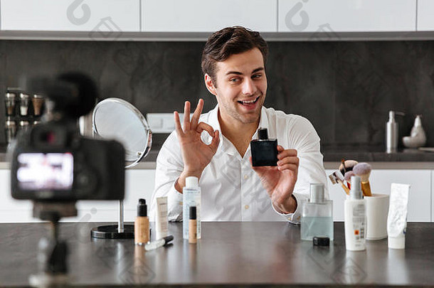 一个快乐的年轻人坐在厨房的桌子旁，一边化妆一边在他的视频博客上拍摄关于新化妆品的插曲