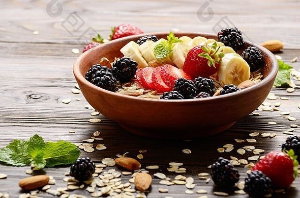 木制厨房餐桌上的粘土盘中有香蕉、草莓、杏仁和黑莓的水果健康慕斯里