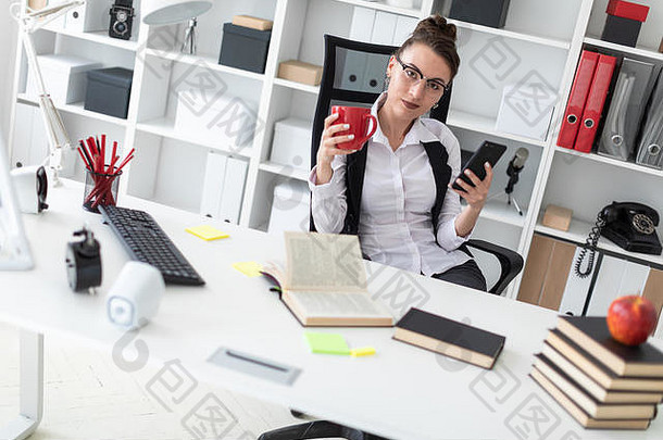 年轻的女孩坐着电脑桌子上办公室持有红色的杯电话