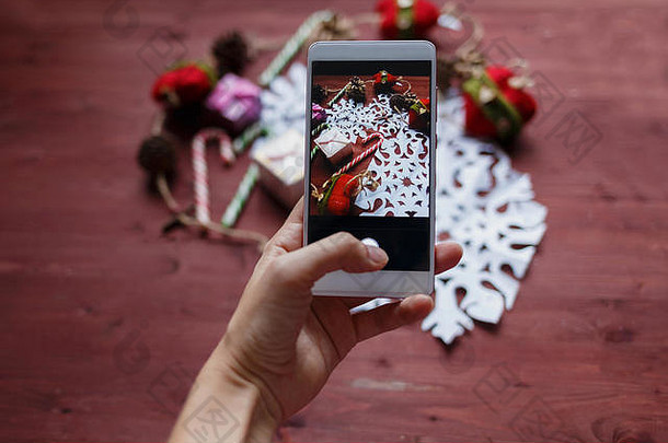 新年的圣诞节概念。女人的手拿着一部智能手机，在木制桌子的复印空间上拍摄从纸上剪下的雪花、礼物、剪刀