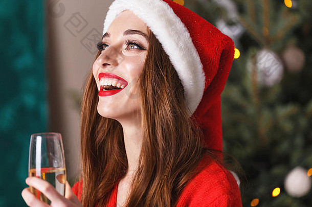 在新年、新年、圣诞节、假期、纪念品、礼品、<strong>购</strong>物、折扣、商店、雪人圣诞老人、化妆、发型、<strong>狂欢</strong>节前几分钟，穿着圣诞帽和红色毛衣的美丽圣诞妇女在舒适的新年云杉旁喝香槟的肖像。