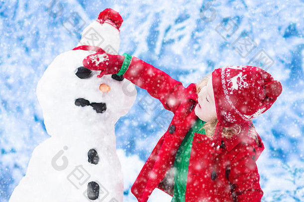 一个滑稽的蹒跚学步的小女孩，戴着一顶红色的北欧针织帽子和一件暖和的外套，和一个雪人玩耍。冬天孩子们在户外玩耍。