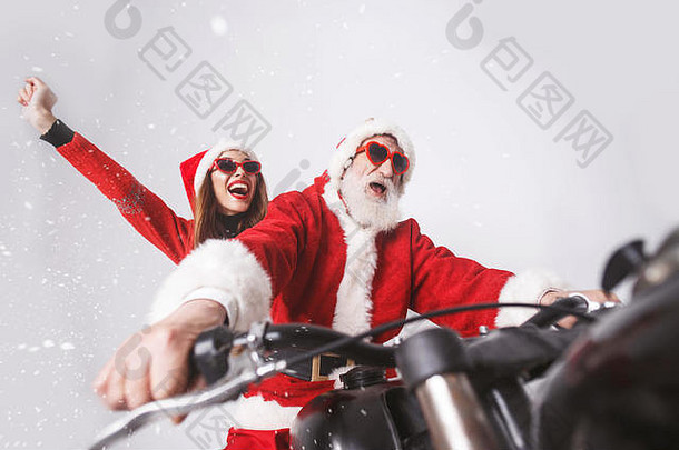 雪地里，白胡子的圣诞老人和戴着圣诞帽、红色毛衣和太阳镜的年轻老太太骑着摩托车在雪地里欢呼雀跃。新年、圣诞节、节假日、纪念品、礼物、<strong>购</strong>物、折扣、商店、雪人圣诞老人、化妆、发型、<strong>狂欢</strong>节。