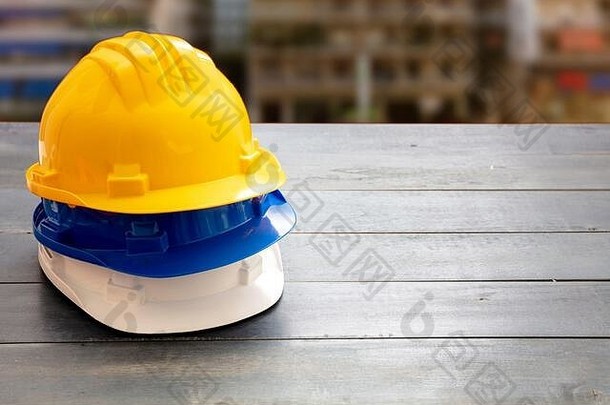 安全生产防护设备。工业防护安全帽白色、黄色和蓝色堆放在木架上，模糊施工现场背景