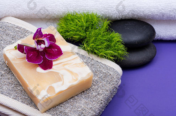 Spa健康概念。天然棉和亚麻籽后洗涤器，杏仁山羊奶皂，玄武岩石头，兰花和石竹花紫色背景。