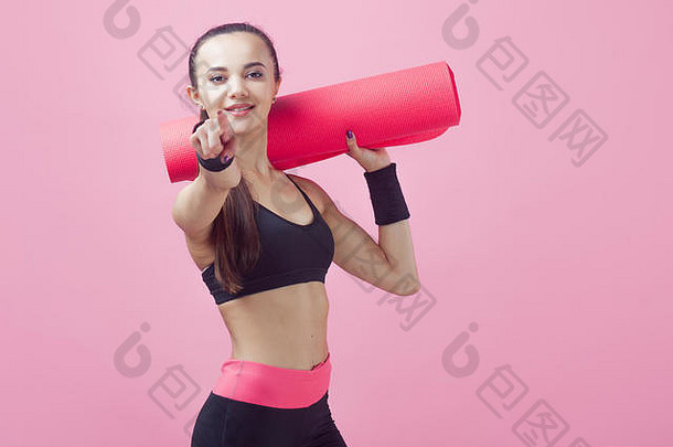 一个黑发女孩，迷人，站在粉红色的背景上，胳膊下夹着<strong>瑜伽垫</strong>，她的左手用手指指向相框。