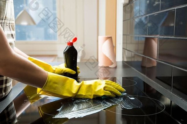 家庭主妇用橡胶防护清洁和抛光电饭锅。厨房顶部黑色闪亮表面，手上有泡沫、海绵、一瓶清洁剂