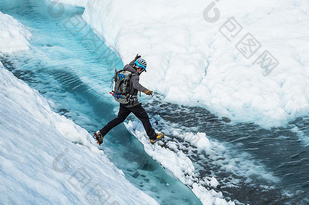 阿拉斯加州马塔努斯卡冰川上，年轻的攀冰向导在一个充满蓝色水的裂缝上跳跃。