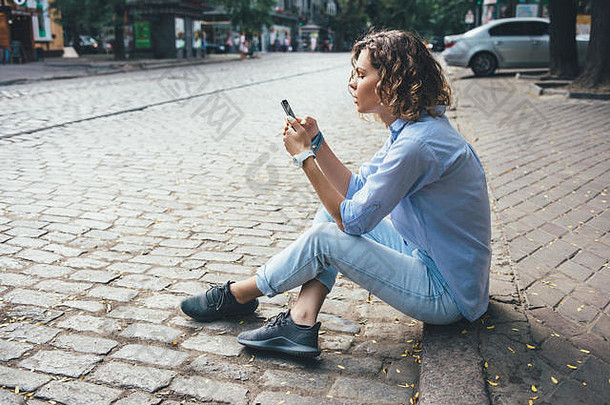 <strong>时尚休闲</strong>的年轻女子坐在路边的人行道上使用智能手机。穿着蓝色衬衫、牛仔裤和<strong>运动</strong>鞋的女旅行者