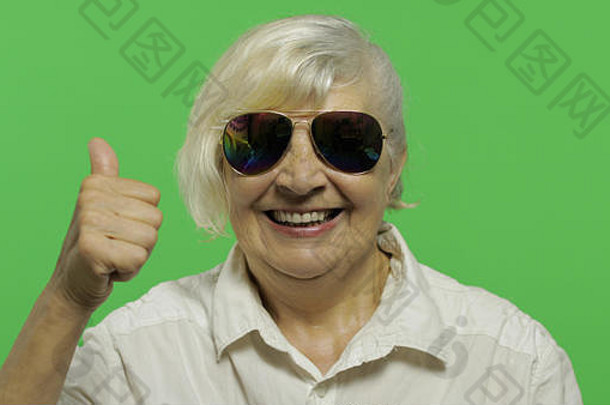 一位戴太阳镜的老妇人竖起大拇指微笑着。穿着白衬衫的漂亮老奶奶。放置您的徽标或文字。色度键。绿色屏幕背景