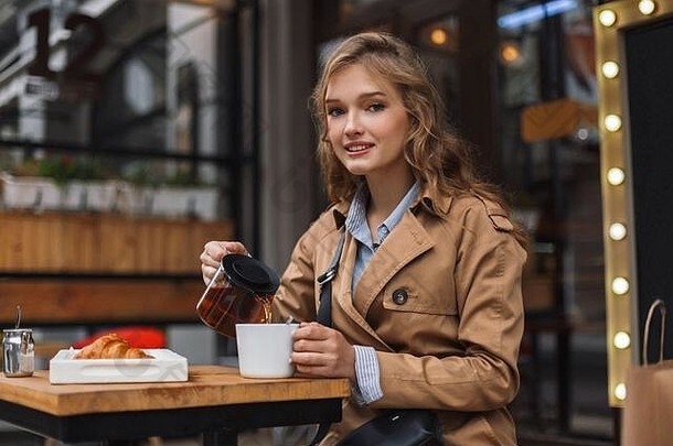 穿着风衣的年轻漂亮女人在舒适的咖啡馆露台上拿着茶壶和白杯子，梦幻般地看着镜头