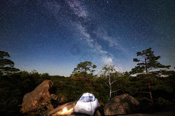 夏夜在落基山脉露营。照亮了游客的帐篷和篝火，在充满星星和银河的令人惊叹的夜空<strong>下</strong>。背景是美丽的<strong>星空</strong>、大石头和树木