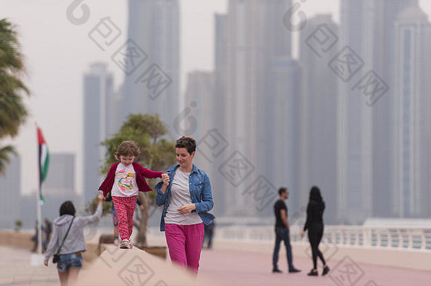 年轻的母亲和可爱的小女孩在海边的长廊上欢快地奔跑，背景是一座大城市