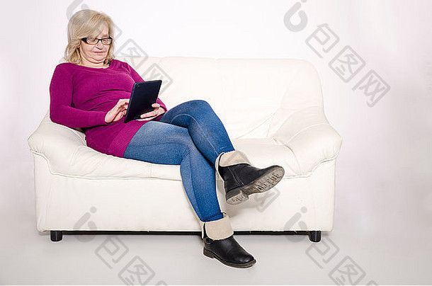坐在白色沙发上，在平板电脑上工作的老妇人。