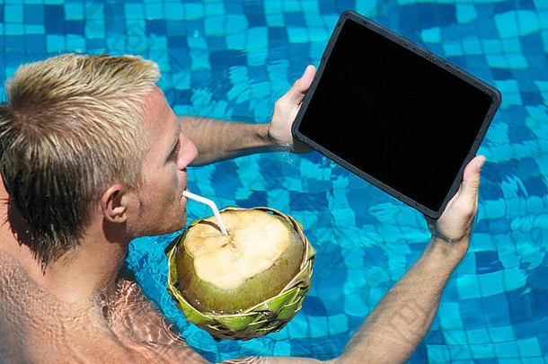 年轻人在热带游泳池喝椰子水鸡尾酒时，一边用平板电脑放松