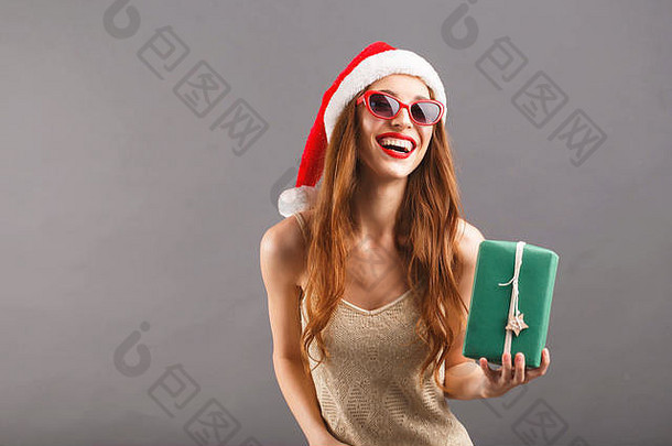 戴着红色圣诞老人帽子和红唇太阳镜的滑稽漂亮女人拿着绿色包装的礼物，她笑着站在灰色背景上，新年、圣诞节、假期、纪念品、礼物、<strong>购</strong>物、折扣、商店、雪人圣诞老人、化妆、发型、<strong>狂欢</strong>节。