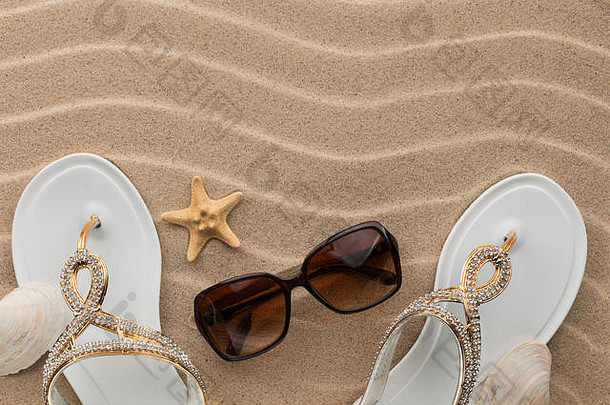 白色的人字拖，有水钻和太阳镜，贝壳，海星在波浪状的沙滩上。为您的文本添加位置
