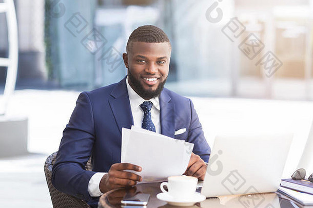 黑人商人在户外咖啡馆拿着纸和笔记本电脑工作