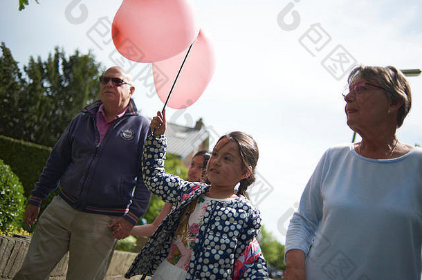 祖父母和他们的孙女在街上散步，孙女在她的生日那天举着一个心形的大气球