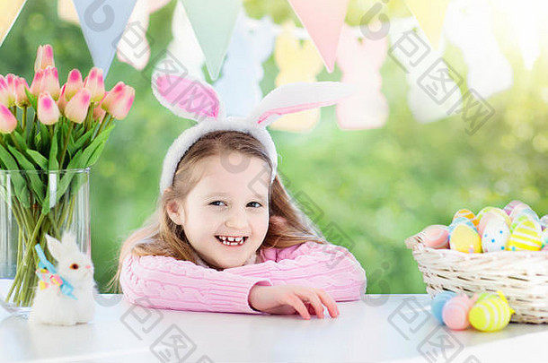 复活节早晨早餐时，穿着兔子耳朵的滑稽小女孩坐在桌子旁，手里拿着复活节彩蛋篮。孩子们庆祝复活节。孩子们在寻找复活节彩蛋。