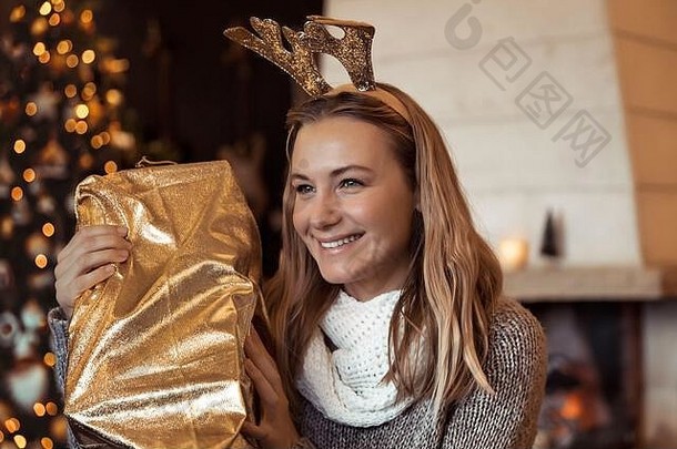 在装饰好的圣诞树旁，一个女孩的画像，她拿着大礼物，戴着可爱的驯鹿角，圣诞快乐，新年快乐