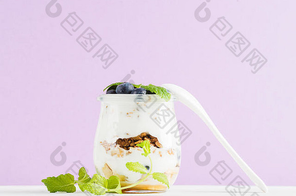 酸奶燕麦格兰诺拉麦片，配果酱、蓝莓和绿叶，置于淡紫色背景下的玻璃罐中。