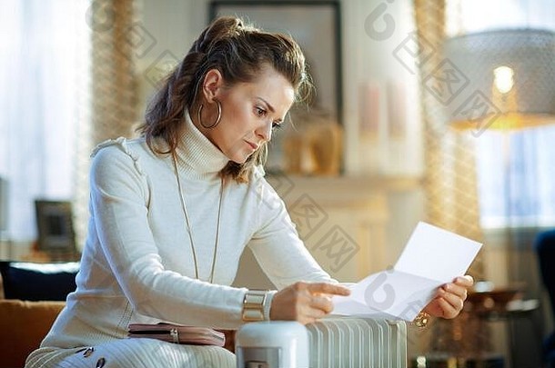 在美国南部的现代客厅里，穿着白色毛衣和裙子的不开心的时髦家庭主妇坐在沙发上，拿着公用事业账单，旁边是白色的电热油散热器