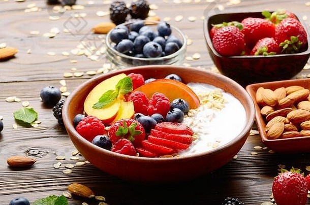 在木制厨房餐桌上，用粘土盘和酸奶，配上桃子、草莓、杏仁和黑莓，水果健康的慕斯利