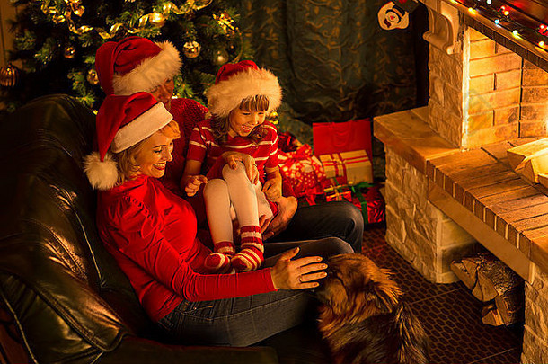 戴着红帽子的幸福家庭，可爱的小狗坐在壁炉旁的圣诞树旁