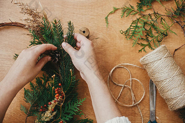 手拿着杉木<strong>树</strong>枝和松果、浆果、线、剪刀放在木桌上。制作质朴的圣诞花环。工作坊详情