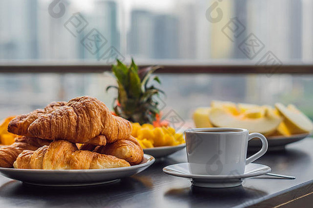 在大城市背景下的阳台上，早餐桌上摆着咖啡、水果和面包